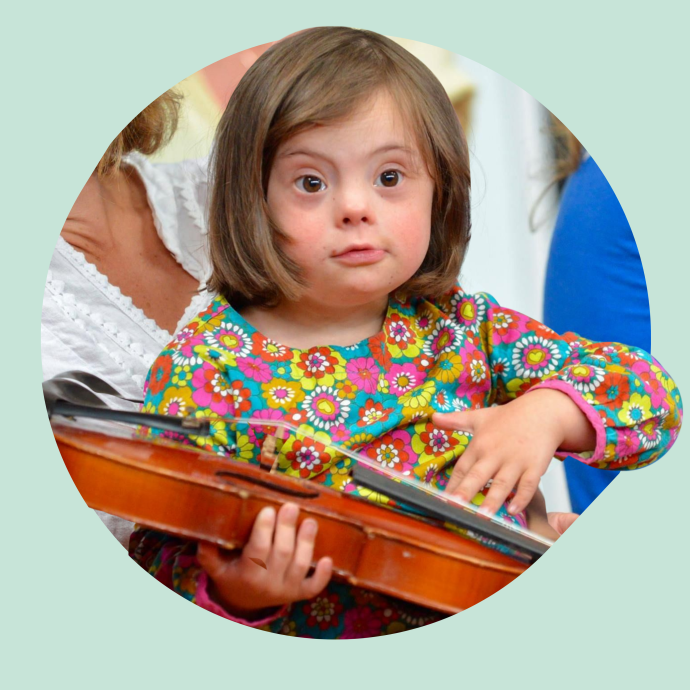 A child holding a violin, wearing a colorful floral-patterned shirt, with an adult partially visible in the background assisting.